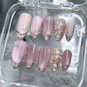Op de afbeelding: Een set van tien kunstnagels in een doorzichtig plastic doosje. De nagels hebben een mauve kattenoog-afwerking met metallic goudkleurige, verhoogde lijndetails op de punten. Twee van de nagels tonen een wit bloemmotief met gouden omlijningen.