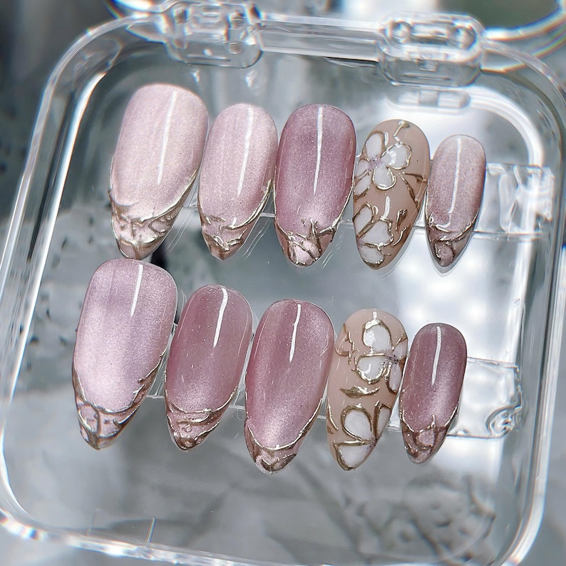 Op de afbeelding: Een set van tien kunstnagels in een doorzichtig plastic doosje. De nagels hebben een mauve kattenoog-afwerking met metallic goudkleurige, verhoogde lijndetails op de punten. Twee van de nagels tonen een wit bloemmotief met gouden omlijningen.