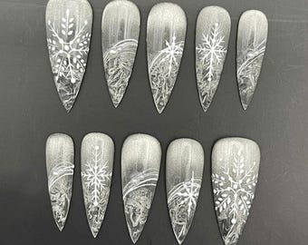 Puede incluir: Un juego de diez uñas postizas en forma de estilete con una base de degradado de purpurina plateada, diseños de copos de nieve pintados a mano en blanco, líneas blancas abstractas y pequeñas cuentas plateadas incrustadas.