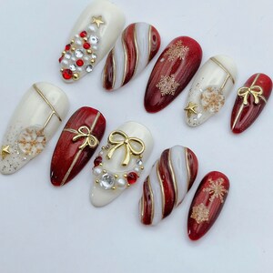 May include: A set of ten press-on nails featuring a holiday theme with red and white color palettes. The designs include gold bows, metallic snowflakes, swirling candy cane patterns, and embellishments of small white pearls and red crystals.