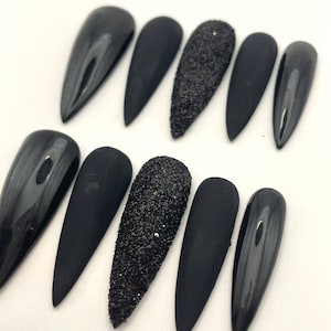 May include: A set of ten stiletto-shaped press-on nails in black. The collection features a mix of finishes, including glossy black, matte black, and textured black glitter nails, arranged in two rows of five on a plain white surface.