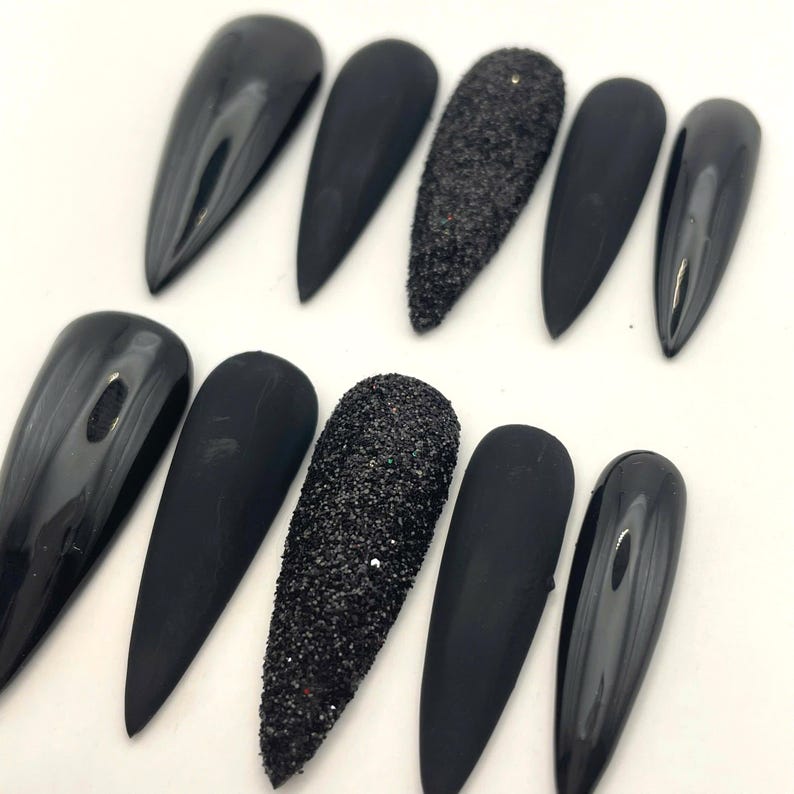 May include: A set of ten stiletto-shaped press-on nails in black. The collection features a mix of finishes, including glossy black, matte black, and textured black glitter nails, arranged in two rows of five on a plain white surface.