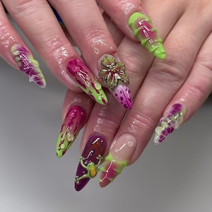 May include: Long almond-shaped nails featuring a vibrant mix of lime green and magenta polish. The design includes three-dimensional elements such as a small green frog figurine, a floral accent, and raised clear droplets across the nail surfaces.