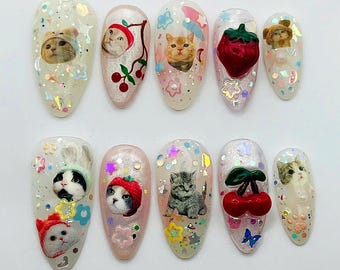 May include: A set of ten almond-shaped press-on nails featuring various designs including cat portraits, red cherries, a red strawberry, and colorful star and flower-shaped glitter accents embedded in a translucent, shimmering base coat.