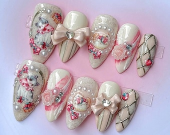 May include: A set of ten almond-shaped press-on nails featuring a cream and beige color palette. The designs include illustrations of kittens, three-dimensional bows with rhinestones, small white pearls, pink roses, and gold lattice patterns.