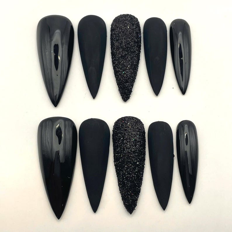 May include: A set of ten stiletto-shaped press-on nails in black. The collection features a mix of finishes, including glossy black, matte black, and a textured black glitter finish on the centre nail of each row, arranged in two horizontal lines.