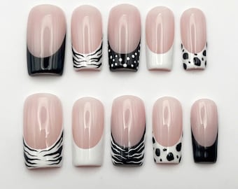 Puede incluir: Un juego de diez uñas postizas con base color nude y varios diseños de puntas en blanco y negro, incluyendo negro sólido, rayas de cebra, puntos blancos, blanco sólido y estampado de vaca, dispuestas en dos filas sobre fondo blanco.