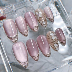 Op de afbeelding: Een set van tien kunstnagels met een mauve kattenoog magnetische afwerking. Sommige nagels zijn effen mauve met zilveren metallic accenten op de punten, terwijl andere witte bloemmotieven hebben omlijnd met zilveren metallic verf op een mauve basis.