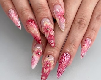 May include: Stiletto-shaped fingernails featuring intricate 3D pink and white floral designs, swirling patterns, and small embedded rhinestones. The nail art incorporates a gradient of deep red to light pink hues with glossy finishes and raised textures.