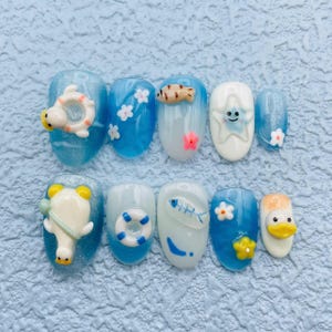 May include: A set of ten press-on nails featuring a summer theme with shades of blue and white. The designs include 3D charms of ducks in life rings, a smiling starfish, small white and pink flowers, a fish skeleton, and a blue life preserver.