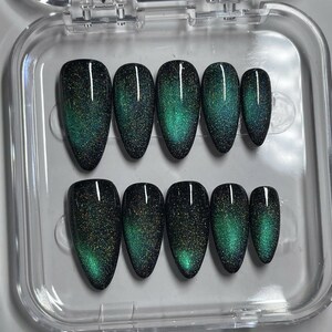 May include: A set of ten almond-shaped press-on nails arranged in a clear plastic storage case. Each nail features a deep black base color with a shimmering, magnetic cat-eye effect that displays a vibrant, glowing emerald green gradient.
