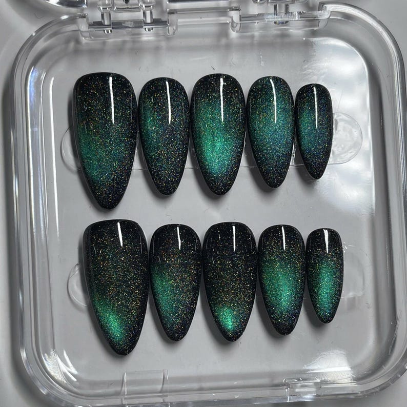 May include: A set of ten almond-shaped press-on nails arranged in a clear plastic storage case. Each nail features a deep black base color with a shimmering, magnetic cat-eye effect that displays a vibrant, glowing emerald green gradient.