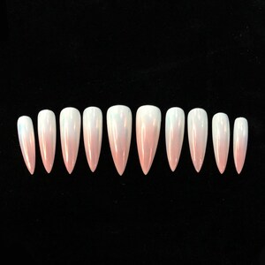 Op de afbeelding: Een set van tien stiletto-vormige kunstnagels met een gladde, parelmoerachtige afwerking die verloopt van zacht lichtroze bij de puntige uiteinden naar doorschijnend wit bij de afgeronde basis, gerangschikt in een horizontale rij op een zwarte achtergrond.