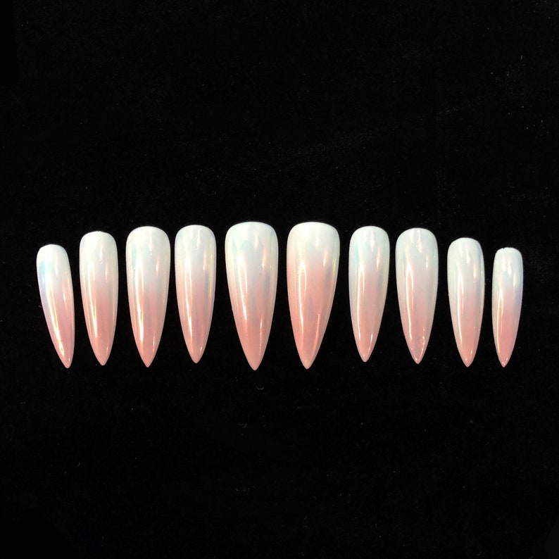 Op de afbeelding: Een set van tien stiletto-vormige kunstnagels met een gladde, parelmoerachtige afwerking die verloopt van zacht lichtroze bij de puntige uiteinden naar doorschijnend wit bij de afgeronde basis, gerangschikt in een horizontale rij op een zwarte achtergrond.