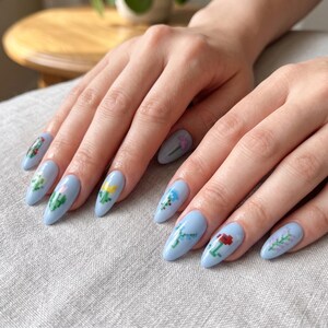 Puede incluir: U&ntilde;as en forma de almendra pintadas con esmalte azul b&iacute;garo claro. Cada u&ntilde;a presenta un peque&ntilde;o dise&ntilde;o floral de arte de p&iacute;xeles distinto en varios colores, incluyendo amarillo, rojo, verde y morado, creando un patr&oacute;n bot&aacute;nico detallado y colorido.