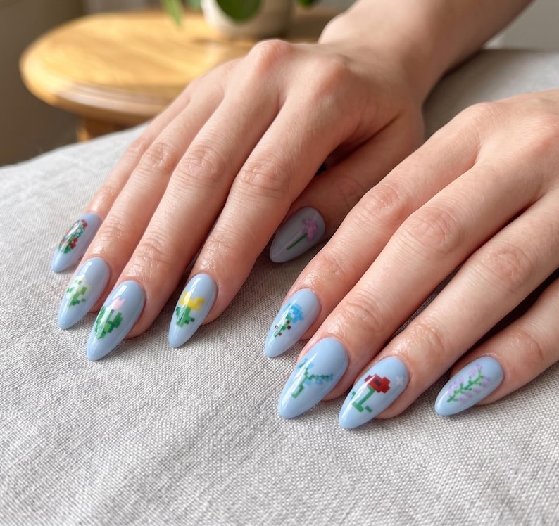 Puede incluir: U&ntilde;as en forma de almendra pintadas con esmalte azul b&iacute;garo claro. Cada u&ntilde;a presenta un peque&ntilde;o dise&ntilde;o floral de arte de p&iacute;xeles distinto en varios colores, incluyendo amarillo, rojo, verde y morado, creando un patr&oacute;n bot&aacute;nico detallado y colorido.