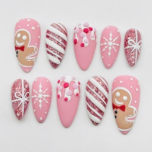 May include: A set of ten almond-shaped press-on nails featuring a pink base with holiday designs including gingerbread figures, white diagonal stripes over pink glitter, white icing drips with pink dots, white snowflakes, and pink glitter gift bows.