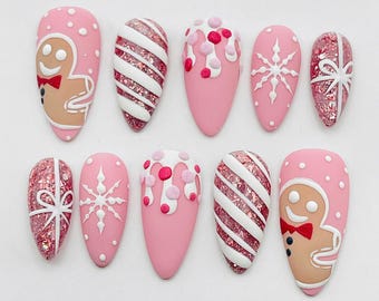 May include: A set of ten almond-shaped press-on nails featuring a pink base with holiday designs including gingerbread figures, white diagonal stripes over pink glitter, white icing drips with pink dots, white snowflakes, and pink glitter gift bows.