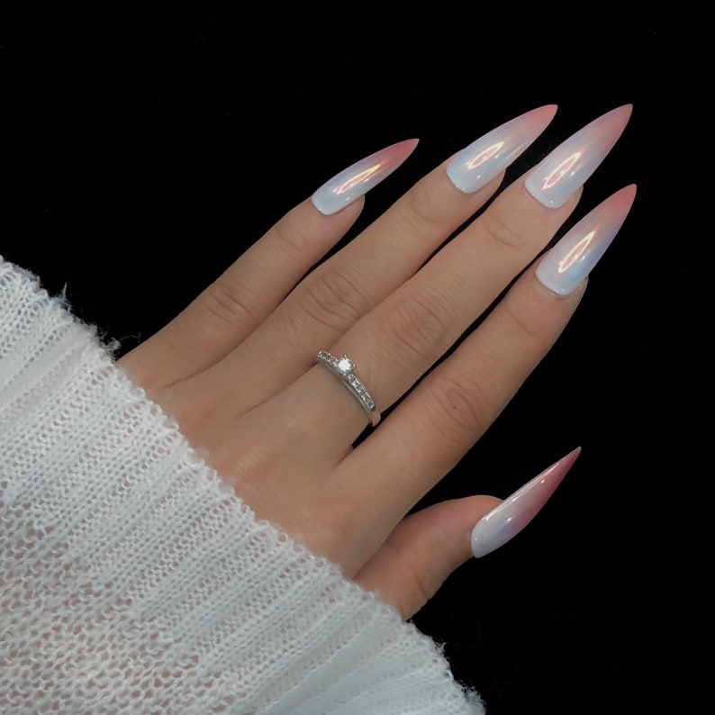 Op de afbeelding: Een zilveren ring met een centrale heldere steen en een band van kleinere steentjes, gedragen aan een vinger met lange, puntige, iriserend witte nagels die naar de punten toe overgaan in een zachte roze tint, tegen een zwarte achtergrond.