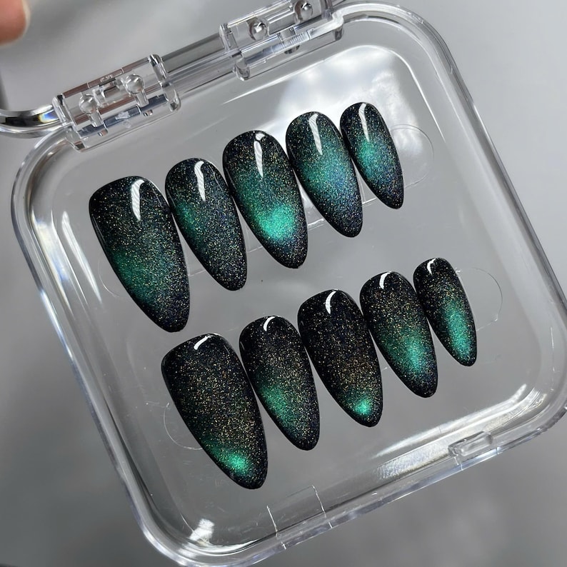 May include: A set of ten almond-shaped press-on nails arranged in a clear plastic storage case. Each nail features a dark black base with a shimmering, magnetic cat-eye effect that displays a vibrant emerald green glow across the surface.