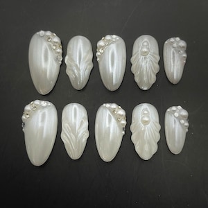 May include: A set of ten pearlescent white artificial nails. Some nails feature rhinestone and pearl embellishments. The nails have an oval shape and are arranged on a dark surface.