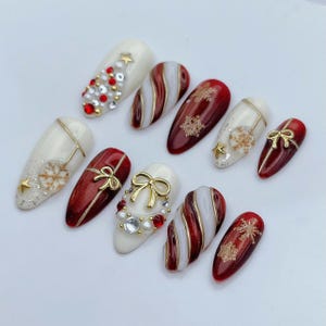 May include: A set of ten almond-shaped press-on nails featuring a Christmas theme. The designs include red and white candy cane stripes, gold snowflake accents, small gold bows, white pearls, red rhinestones, and gold star charms on glossy surfaces.