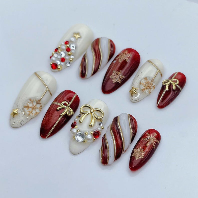May include: A set of ten almond-shaped press-on nails featuring a Christmas theme. The designs include red and white candy cane stripes, gold snowflake accents, small gold bows, white pearls, red rhinestones, and gold star charms on glossy surfaces.
