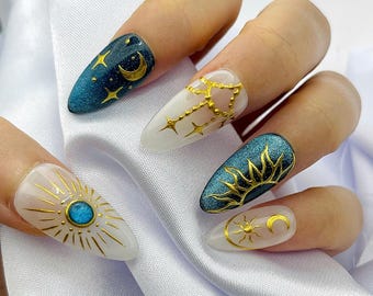 May include: Almond-shaped press-on nails featuring a celestial theme. Two nails are deep teal with gold crescent moon and sun ray designs. Two nails are translucent white with gold star constellations and a central blue stone with sun rays.