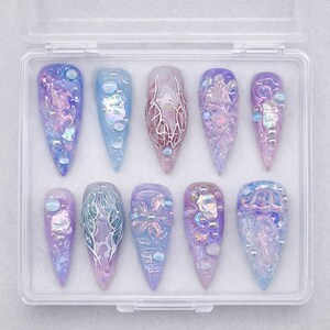 May include: A set of ten almond-shaped press-on nails in a clear plastic storage case. The nails feature a color palette of lavender, periwinkle, and light blue, decorated with iridescent flakes, raised gel textures, and small spherical beads.