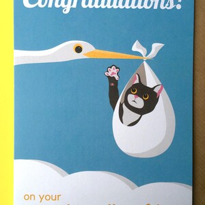 Op de afbeelding: Wenskaart met een ooievaar die een babykat in een eivormige bundel draagt. De kaart heeft de tekst "Congratulations!" en "on your new bundle of joy" in witte en oranje letters op een blauwe achtergrond.