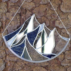 Pu&ograve; includere: Un acchiappasole in vetro colorato a forma di semicerchio con un design a catena montuosa. L'oggetto &egrave; composto da segmenti di vetro blu scuro, testurizzato e a specchio riflettente, uniti da linee di saldatura in metallo argentato e appeso a una catena.
