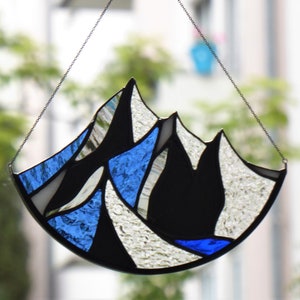 Pu&ograve; includere: Un acchiappasole in vetro colorato con un design a catena montuosa, composto da segmenti di vetro blu, bianco e nero testurizzati uniti da linee di saldatura in metallo scuro, appeso a una sottile catena metallica fissata a un gancio adesivo.