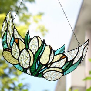 May include: A stained glass crescent moon suncatcher with a floral design. The piece features yellow, green, and clear textured glass, with a black chain for hanging. The design includes tulip-like shapes.