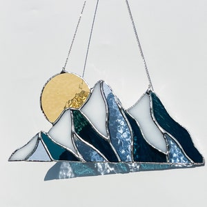 May include: A stained glass sun and mountain scene hanging from a silver chain. The sun is yellow and the mountains are blue, white, and green.