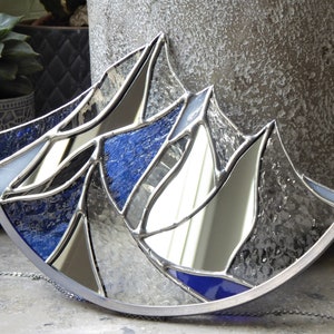 Pu&ograve; includere: Un acchiappasole in vetro colorato semicircolare con un design a catena montuosa. L'oggetto &egrave; composto da pannelli di vetro trasparente, testurizzato e blu profondo uniti da linee di saldatura in metallo argentato, con una sottile catena argentata.