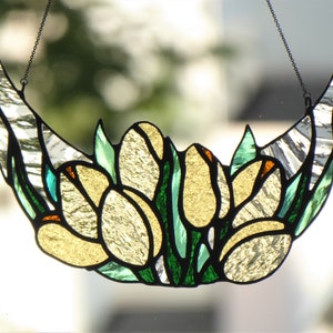 May include: Stained glass crescent moon with yellow tulip-like flowers and green leaves. The glass has a textured, iridescent quality, with black outlines and a chain for hanging. The design is reminiscent of Art Nouveau.