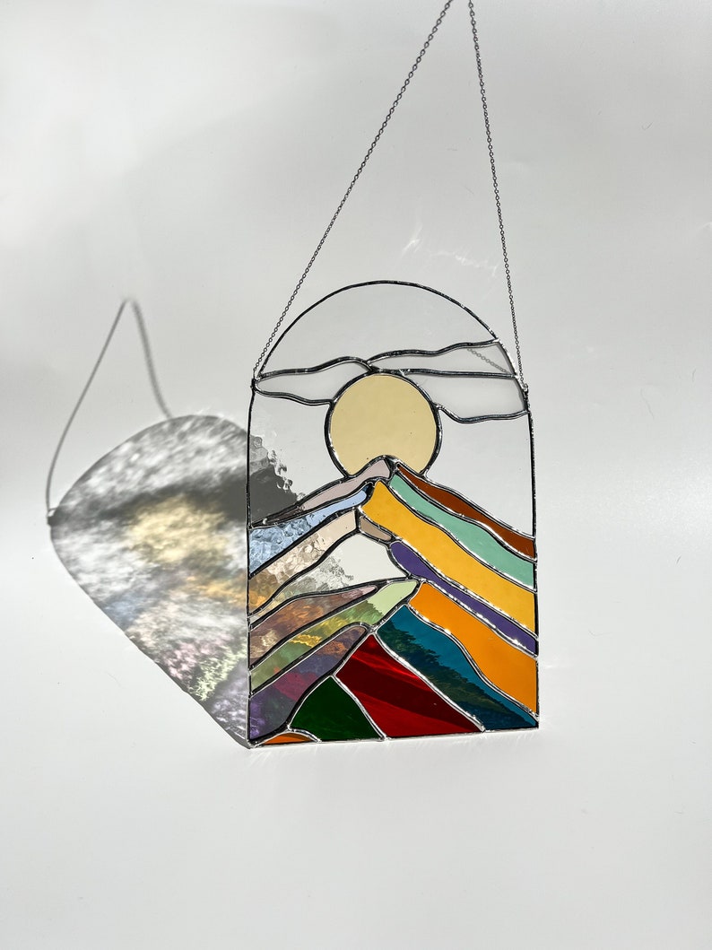 May include: A stained glass window hanging with a chain. The window depicts a colorful mountain range with a yellow sun and clouds in the sky.