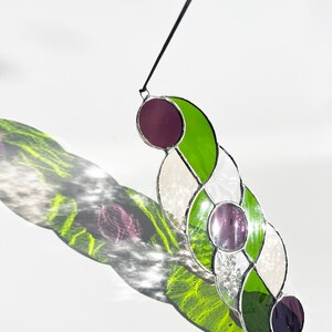 Puede incluir: Un atrapasoles de vidriera con una serie de paneles ovalados conectados. Los paneles presentan tonos de vidrio verde, blanco y morado, con un cord&oacute;n negro para colgar. El atrapasoles proyecta una colorida sombra sobre la superficie blanca.