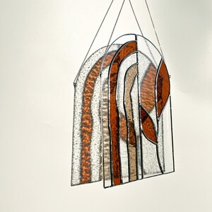 May include: Stained glass window panel with an arched design. The panel features a mix of clear, amber, and brown glass pieces, creating a warm aesthetic. The glass is held together by black metal framing, and it hangs from a silver chain.