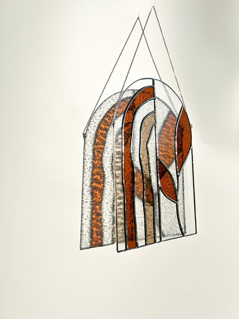May include: Stained glass window panel with an arched design. The panel features a mix of clear, amber, and brown glass pieces, creating a warm aesthetic. The glass is held together by black metal framing, and it hangs from a silver chain.