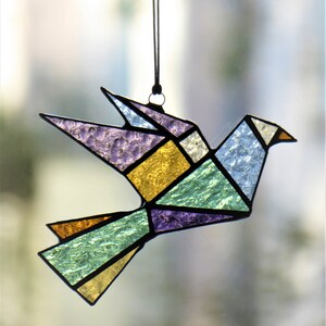 Pode incluir: Um ornamento de pomba em vitral com um design geom&eacute;trico. O p&aacute;ssaro apresenta v&aacute;rias cores, incluindo roxo, amarelo, verde e azul, com contornos pretos. Ele pende de uma corda preta, adequado para exibi&ccedil;&atilde;o.