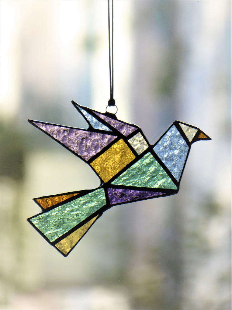 Pode incluir: Um ornamento de pomba em vitral com um design geom&eacute;trico. O p&aacute;ssaro apresenta v&aacute;rias cores, incluindo roxo, amarelo, verde e azul, com contornos pretos. Ele pende de uma corda preta, adequado para exibi&ccedil;&atilde;o.