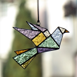 Pode incluir: Um ornamento de pomba em vitral com um design geom&eacute;trico. O p&aacute;ssaro apresenta v&aacute;rias cores, incluindo roxo, dourado, verde e azul, com contornos pretos. Ele pende de um cord&atilde;o preto, adequado para decora&ccedil;&atilde;o de janelas ou paredes.