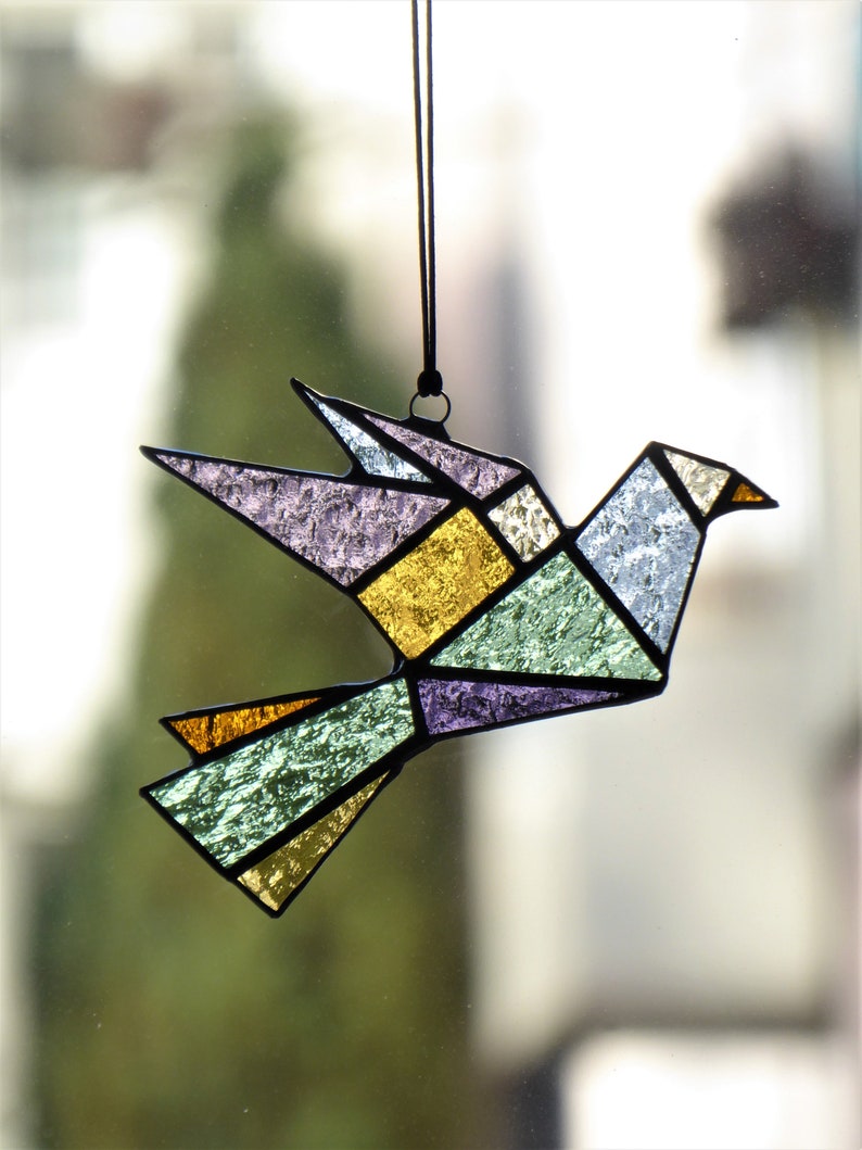 Pode incluir: Um ornamento de pomba em vitral com um design geom&eacute;trico. O p&aacute;ssaro apresenta v&aacute;rias cores, incluindo roxo, dourado, verde e azul, com contornos pretos. Ele pende de um cord&atilde;o preto, adequado para decora&ccedil;&atilde;o de janelas ou paredes.