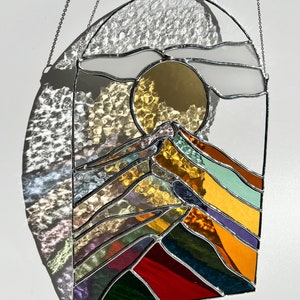 May include: A stained glass window hanging with a chain, depicting a stylized mountain range with a sun and clouds in the background. The mountains are made of colorful glass, including red, orange, yellow, green, blue, and purple.