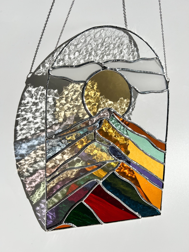May include: A stained glass window hanging with a chain, depicting a stylized mountain range with a sun and clouds in the background. The mountains are made of colorful glass, including red, orange, yellow, green, blue, and purple.