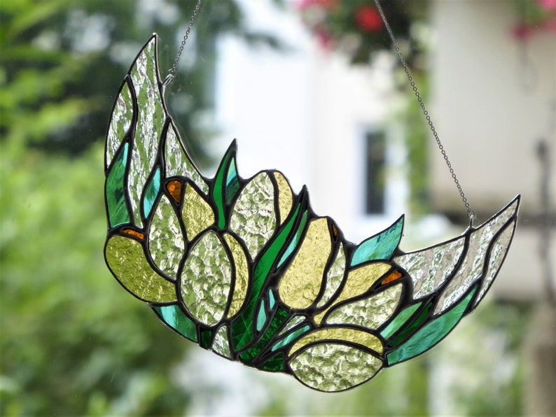 May include: Stained glass crescent moon with a floral design. The artwork features green, yellow, and clear textured glass, with black lines defining the shapes. A chain is attached for hanging. The design includes tulip-like flowers.
