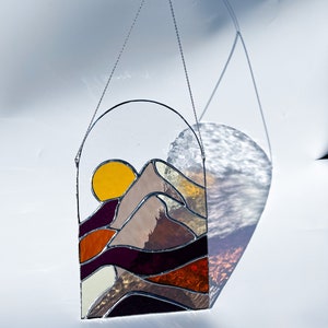 K&ouml;nnte beinhalten: Ein Buntglasfenster, das an einer Kette h&auml;ngt. Das Fenster zeigt einen Sonnenuntergang &uuml;ber einem Gebirge mit einer gelben Sonne, orangefarbenen, braunen und violetten Bergen.