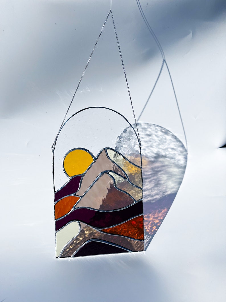K&ouml;nnte beinhalten: Ein Buntglasfenster, das an einer Kette h&auml;ngt. Das Fenster zeigt einen Sonnenuntergang &uuml;ber einem Gebirge mit einer gelben Sonne, orangefarbenen, braunen und violetten Bergen.