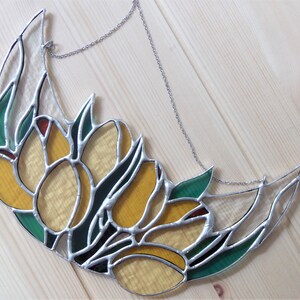 May include: Stained glass crescent moon with yellow tulip flowers, green leaves, and red accents. The glass is outlined with silver solder and hangs from a silver chain. The artwork is set against a light wood background.